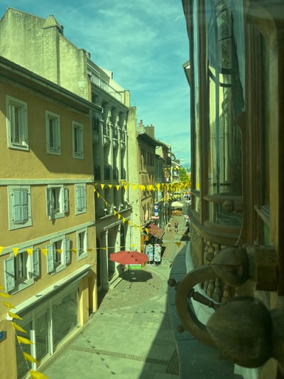 The image shows a street scene in Évian-les-Bains, France, viewed from an elevated perspective, likely a balcony or window. The perspective is from the right side of the frame, looking down a narrow cobblestone street lined with buildings. The buildings are predominantly light yellow and white, with several stories and windows. Yellow bunting flags are strung across the street, creating a festive atmosphere.
On the left side of the street, a yellow building with multiple windows and shutters is prominently displayed. Some windows are open, revealing glimpses of interiors. A storefront with a large glass window is visible on the ground floor.
Across the street, the architecture is more varied. There are buildings with columns and balconies. Several shops with awnings are visible, and a red umbrella stands open in front of one. A few people are walking along the street, some appearing to be shoppers or tourists. The street itself is paved with cobblestones in some areas and a lighter colored stone in others.
The sky is partly cloudy with a light blue hue. In the distance, mountains can be seen. The right side of the image is dominated by a large, curved window with decorative wooden framing and what appears to be a wooden ship's wheel detail. Reflections are visible in the glass, obscuring some of the interior details. The overall lighting suggests it is daytime, possibly late morning or early afternoon. The green tint of the image may be due to the lighting conditions or a color filter.