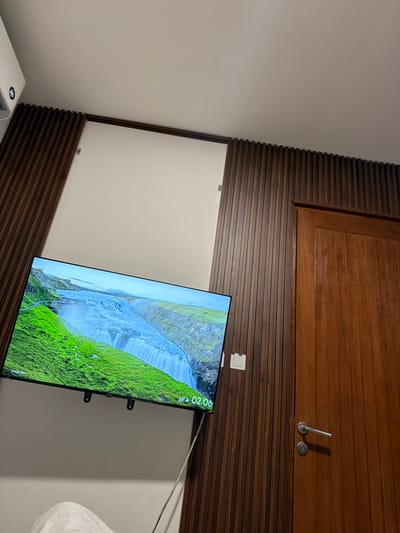 The image shows a close-up of a modern living room setup with a focus on a large flat-screen television mounted on a wall. The TV displays a scenic landscape of a waterfall cascading down a lush green hillside, with a partially visible digital clock showing "02:06" and a temperature of "26°". The wall behind the television features a distinctive horizontal wooden paneling design, with a larger wooden cabinet or panel above it. Two small silver hooks or brackets are visible on the wooden paneling. The overall ambiance is modern and minimalist, with a neutral-colored wall contrasting with the rich wood tones. The scene appears to be indoors, likely in a residential setting, given the presence of the TV and the overall home decor. The lighting suggests it could be daytime or evening, with ambient light reflecting off the screen and surfaces. No people are visible in the frame.