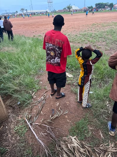 This image depicts an outdoor scene on a dirt field in Du, Nigeria, likely during a soccer match. The foreground features two young boys. The boy on the left is wearing a red t-shirt with a graphic design and black shorts. The boy on the right is wearing a colorful, patterned long-sleeved shirt and pants. Both boys are standing on a dirt path partially covered with dry leaves and grass. In the background, a group of people, some of whom appear to be players, are engaged in a soccer game on a red dirt field under an overcast sky. Several buildings and trees are visible in the distance. The overall environment suggests a casual sporting event in a rural or semi-urban setting.