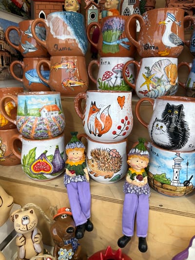 The image shows a display of handcrafted ceramic mugs and figurines, likely in a souvenir shop. The items are arranged on wooden shelves. Many of the mugs are decorated with painted scenes and motifs. Some feature marine life like dolphins, fish, and seashells, while others depict pastoral landscapes with houses and vineyards, or whimsical designs with mushrooms, foxes, and cats. The word "Ялта" is visible on several of the mugs, indicating the location is Yalta, Ukraine. There are also several small, doll-like figurines of children, one with a hedgehog, and other decorative ceramic pieces like a cat and a dog. The lighting suggests an indoor setting, possibly with natural light coming from the side or front.