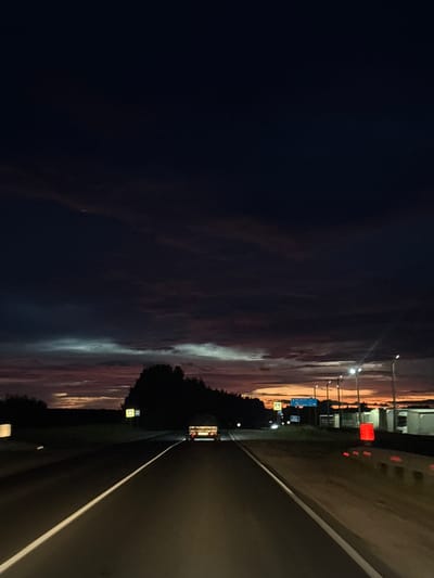 This media file depicts a nighttime scene on a highway in Kotelnich, Russia. The image is oriented vertically. The left side of the frame is dominated by a dark, almost black sky with some faint, wispy clouds that show subtle hues of purple and orange from the distant sunset or twilight. Streetlights are visible along the right side of the highway, casting pools of light.
The highway itself occupies the right side of the image, with a clear white dividing line. The road surface appears dark and wet, suggesting recent rain or dew. A vehicle, possibly a van or truck, is seen driving away in the distance on the highway, its taillights illuminated. Along the side of the road, there are various signs and structures. One prominent sign is blue with white text, and another is a yellow square sign with a black symbol inside. There are also red lights visible on what appears to be a barrier or structure along the roadside. Dark trees form a silhouette along the edge of the highway, further emphasizing the nighttime setting. The overall mood is one of a solitary journey at dusk or early night. The sky's colors suggest the time is either very late evening or very early morning.