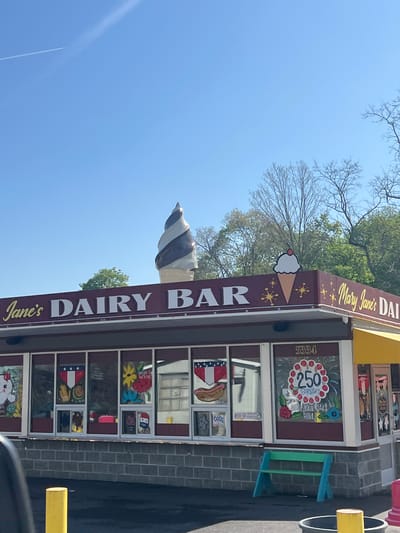 The image, rotated, depicts the vibrant exterior of Mary Jane's Dairy Bar under a clear blue sky. A large, realistic sculpture of a chocolate and vanilla swirl ice cream cone adorns the roof, alongside a smaller, flat ice cream cone sign topped with a cherry. The building's windows are extensively decorated with colorful paintings, including an American flag motif, a hot dog, various flowers, and cartoon figures. One prominent window features a large birthday cake design with "250 BIRTHDAY YOUR CAKE HERE" visible, indicating a special event or offer. The address "5334" is also displayed above a window. Other visible text includes "GOD" and "PICK UP C" on window decals. In front of the establishment, a small blue and green picnic bench is present, along with yellow bollards. The background shows bare tree branches, suggesting a seasonal transition, interspersed with some green foliage. A faint airplane contrail streaks across the bright sky. No people are visible, and the scene depicts a sunny day.