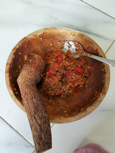 A wooden bowl contains a vibrant red chili paste. A spoon rests on top of the paste. A wooden pestle leans against the inside of the bowl. The bowl's interior reveals a dark, stained wood. The background is a light-colored tiled surface. A corner of a pink object is visible in the bottom right.