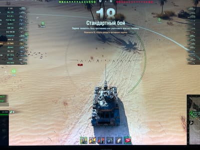 The image is a screenshot from a video game, likely a tank combat simulator like World of Tanks. The perspective is from a third-person view behind a damaged tank on a desert map. The game interface displays various elements including a minimap in the bottom right corner, a list of tanks on the left side, ammunition and equipment slots at the bottom, and in-game text and indicators in the center of the screen. The central text reads "Стандартный бой" (Standard Battle) and "Задача: захватить базу противника или уничтожить всю его технику" (Objective: capture the enemy base or destroy all their equipment). Below this, it says "Нажмите N, чтобы увидеть активные задачи" (Press N to see active missions). A timer shows "17:01". There is no indication of the city of Grozny, Russia; the environment is clearly a desert with sand dunes and sparse vegetation. The only identifiable object besides the tanks is a tank itself in the foreground, appearing heavily damaged and with a burnt texture.