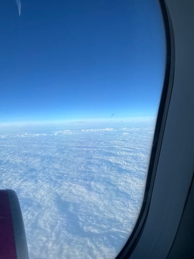 An aerial photograph, taken from inside an aircraft, depicts a high-altitude view over or in the vicinity of Caselle Torinese, Italy. The upper two-thirds of the image display a clear, deep blue sky, gradually lightening towards the horizon. Below this, a vast, unbroken expanse of white, textured cloud cover extends horizontally, obscuring all ground features. The right side of the frame is defined by the dark gray and black molding of an aircraft window. A curved, magenta-colored structural component, consistent with a portion of an aircraft engine nacelle or wing, is visible in the bottom-left quadrant. Minor specks and smudges are present on the window surface. No individuals, specific actions, or interactions are visible within the scene.