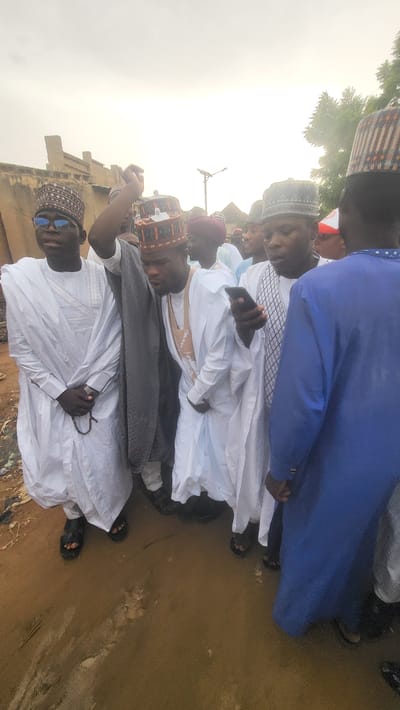 A group of men, primarily young to middle-aged adults, are gathered outdoors in Jogana, Nigeria. Most are dressed in traditional West African attire, including long white robes and various patterned prayer caps (kufis). One man in the foreground wears sunglasses and holds a string of prayer beads in his left hand. Another individual, partially visible, wears a vibrant blue robe. Several men are seen holding mobile phones.

The setting appears to be an open outdoor space, possibly a street or unpaved area. The ground is visibly muddy and wet, indicating recent rainfall or standing water. In the background, partially obscured by a bright, almost washed-out sky, are some low-rise buildings and a street light pole. The bright sky suggests it is daytime, possibly early morning or late afternoon with intense light, or under hazy conditions. The men are standing and looking in various directions, some seemingly observing their surroundings or engaging with others in the group. There is no specific, overt activity such as a ceremony or work, but rather a casual gathering. No visible text can be identified in the image.