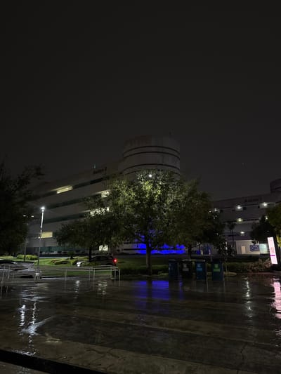 At night in Heroica Puebla de Zaragoza, Mexico, a tree illuminated by streetlights stands in front of the modern white building complex "Aulas 2." The ground appears wet, reflecting blue light from a nearby source. Trash cans and racks are visible along the right edge of the frame. The sky is dark, suggesting overcast conditions.