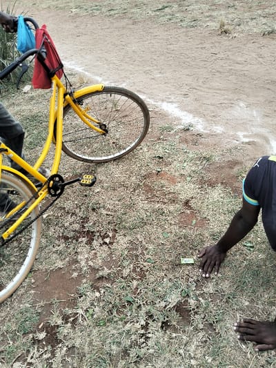 The image depicts an outdoor scene in Waa Ngombeni ward, Kenya. The ground surface consists of reddish-brown soil with sparse patches of dry, light green and brown grass. A distinct white line is visible on the dirt ground in the upper right portion of the frame, extending horizontally.
A bright yellow bicycle is partially present on the left side, angled with its front wheel facing the upper right. A blue plastic bag and a red fabric bag are suspended from the black handlebars. The bicycle has yellow mudguards, a visible chain, and a black pedal with yellow accents. The lower legs of an individual, wearing dark trousers, are visible next to the bicycle's frame on the far left.
On the right side of the frame, another individual is partially visible, appearing to be kneeling or crouching. This person wears a dark blue shirt with light blue and yellow trim on the right sleeve. Their dark-skinned hands are placed palms down on the ground. Directly in front of their hands, a small, rectangular, green object rests on the soil amidst the grass.