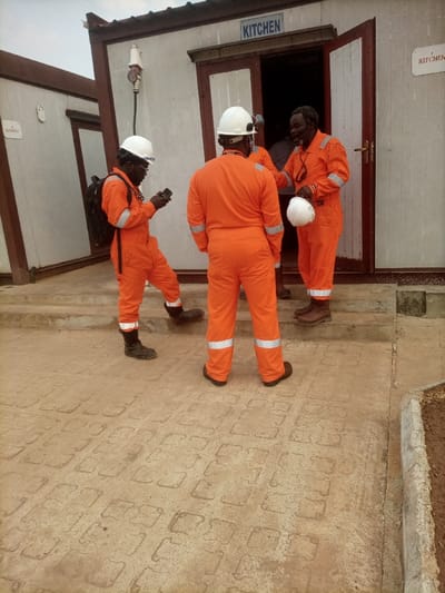The scene, located in Opete, Nigeria, depicts three individuals, identified as black males, wearing orange work coveralls with reflective stripes and white hard hats, standing on a paved area outside two modular buildings under an overcast sky.
The primary building on the right is light-colored with brown trim and has a partially open brown door. A blue sign with white text above this door reads "KITCHEN." A smaller, identical "KITCHEN" sign is present further right on the building's facade. An electrical outlet with a red plug is mounted on the exterior wall to the left of the door.
On the left side of the frame, one male subject wears a black backpack and black safety boots. He holds and views a black smartphone with both hands, with his right foot raised onto a concrete step leading to the "KITCHEN" building. A second male subject stands centrally with his back to the camera, facing the "KITCHEN" doorway. The third male subject, an older individual with a beard, stands at the threshold of the "KITCHEN" doorway, holding a white hard hat in his left hand, with dark glasses hanging from his neck. He is facing the central figure, smiling, indicating a social interaction.
The ground is paved with interlocking light-colored concrete blocks, showing signs of wear and dirt. A concrete step provides access to the "KITCHEN" building entrance. A second modular building, partially visible on the far left, features a sign with text that partially reads "BAKERY." A low concrete retaining wall bordering disturbed earth is visible on the far right.