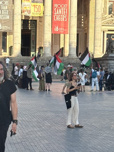 This media file shows a street scene in Brussels, Belgium, with a group of people gathered. Several individuals are holding Palestinian flags, suggesting a protest or demonstration. In the background, there is a building with ornate classical architecture, featuring large columns and decorative carvings. Red banners hang from the building, with visible text in French and English, including "PROOST SANTE! CHEERS" and "150 BELGIAN BEERS". Another banner reads "MORE THAN A BEER MUSEUM". The ground is paved with stone, and the lighting suggests it is daytime, possibly late afternoon. The people in the foreground and middle ground are varied in age and attire, with some wearing casual clothing and others appearing to be part of the demonstration. The overall mood seems to be one of collective action and public gathering, with the flags and banners indicating specific messages or affiliations. One person in the foreground is facing the camera, with a beard and wearing a black t-shirt.