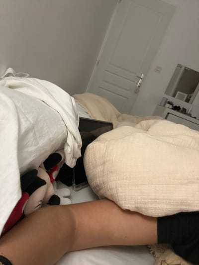 A person lies in bed with a tablet next to them, partially covered by white sheets and a beige blanket. A Mickey Mouse plush toy rests near the tablet. The individual's arm is visible, and they appear to be wearing black clothing. A white door with a latch is in the background. A cabinet or mirror can be seen above the door.