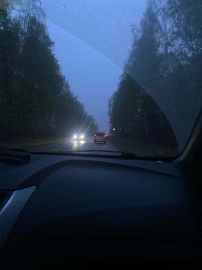 A photograph, taken from inside a vehicle, depicts a road in what appears to be a wooded area. Two vehicles are visible ahead: a car with headlights approaching and a semi-truck with illuminated tail lights further ahead. The view through the windshield is obscured by rain or moisture. The time of day appears to be late evening or early morning, given the subdued lighting. The location is Белоярская Застава, Russia.