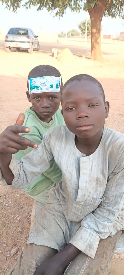 Two dark-skinned young boys are in the foreground. The boy in front is wearing a light, dirty, buttoned shirt and is making a hand gesture. The boy behind him is wearing a green shirt and has a "Sarfi Yogurt" wrapper stuck to his forehead. In the background, there is a light-colored car and a large tree. The ground appears to be dusty or sandy.