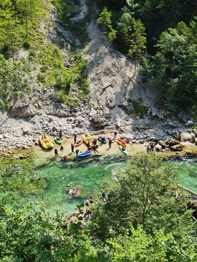 In the immediate foreground, lush green foliage obscures the bottom portion of the image. The midground depicts a group of people and several inflatable rafts and kayaks on the rocky bank of a clear, emerald-green river. The people are wearing life vests and helmets, and some are actively engaged in launching or preparing the watercraft. The riverbank is composed of pebbles and larger stones, with a steep, vegetated slope rising behind it. The background features a dense forest of green trees on a hillside under a bright, sunny sky. This scene is located in the city of Landl, Austria, specifically on the Enns River, known for its white-water rafting and kayaking activities.