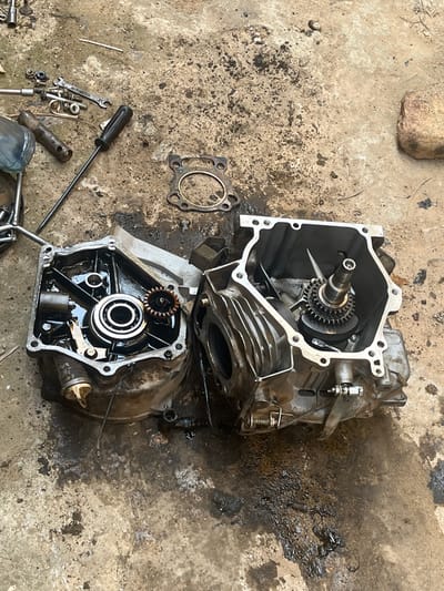 Unguwar Fatika, Nigeria. An engine block is disassembled into two main components on a stained concrete surface. The top half shows a crankshaft with a gear attached. The bottom half shows a main bearing and possibly a camshaft. A gasket, wrenches, and screwdrivers are scattered around the engine parts on the dirty surface.