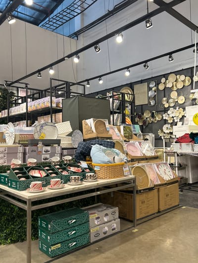 The image depicts the interior of a retail store, likely a home goods or department store, displaying a variety of household items. In the foreground, a metal table is laden with tableware, including patterned cups and saucers, plates, and sets of dishes in green packaging. Behind this table, larger displays showcase woven baskets, decorative wall art, and boxed clocks. Shelving units in the background hold additional merchandise, with boxes and various decorative items visible. The store appears to be well-lit, with track lighting fixtures suspended from the ceiling. The overall impression is one of a retail environment organized for product display and sale, with a wide range of consumer goods. Based on the context provided, the location is a store in Makassar, Indonesia.