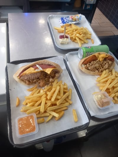 The scene depicts three trays of fast food meals arranged on a stainless steel counter, consistent with a commercial food service establishment in Sallaumines, France.

In the foreground, a black rectangular tray lined with white paper holds an open pita-style sandwich, revealing a patty, melted yellow cheese, a pink deli meat slice, and diced meat. Accompanying this is a generous portion of french fries and a small clear plastic cup of orange-hued sauce. Adjacent, a light gray rectangular tray, also paper-lined, presents another open pita-style sandwich containing diced meat, a breaded item, and melted yellow cheese, alongside french fries, a small clear plastic cup with two distinct sauces (white and light brown), and a horizontal green aluminum beverage can.

In the upper right background, a third light gray, paper-lined tray displays french fries, a small clear plastic cup with white and red sauces and an inserted wooden stick, and a metallic foil packet labeled "Capris."

The immediate environment consists of the stainless steel counter and a dark, possibly tiled, wall in the background. No individuals are present in the frame.