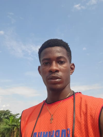 This media file is a portrait-style photograph of a young Black man taken outdoors. He is wearing an orange athletic jersey with blue lettering that is partially obscured. A thin gold chain with a cross pendant is visible around his neck. The man's face is the central focus, with his eyes looking directly at the camera. He appears to be sweating, suggesting warm weather.
The background consists of a bright blue sky with scattered white clouds. To the lower right of the frame, the tops of green leaves, possibly from banana trees or other tropical vegetation, are visible, along with hints of darker foliage. This suggests a location with lush plant life. The lighting and clear sky indicate it is daytime, likely midday or afternoon. The image is oriented sideways.
While the blue lettering on the jersey is partially visible, it is not fully legible. The overall impression is casual and perhaps sporty, given the athletic wear and outdoor setting. The man's expression is neutral or slightly serious. The context provided indicates the location is Egbagu, Nigeria.