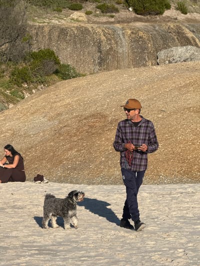 The image captures a scene outdoors, possibly in the vicinity of a rocky landscape near Hout Bay, South Africa. A man in a plaid shirt, dark pants, and a baseball cap stands on a concrete surface with a climbing rope. He is looking at a phone. A shaggy dog lays down next to him. A woman in a brown outfit is visible at the bottom of the frame, towards the corner. The background is dominated by a large rock formation. Vegetation including grass and trees can be seen around the rock.