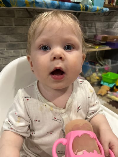 A fair-skinned baby with blonde hair and blue eyes sits in a high chair, looking up with an open mouth. The baby is wearing a light-colored onesie patterned with red hearts and the text "I ❤️ MY FAMILY" and "LOVE." The onesie has some yellow stains on it. The baby is holding a pink sippy cup in their right hand. The background is a cluttered indoor space with toys and other items. A striped blue and gold object is visible to the left of the baby's head. The location appears to be indoors, possibly a kitchen or dining area. No information suggests this image was taken in Vesyoloye, Russia.