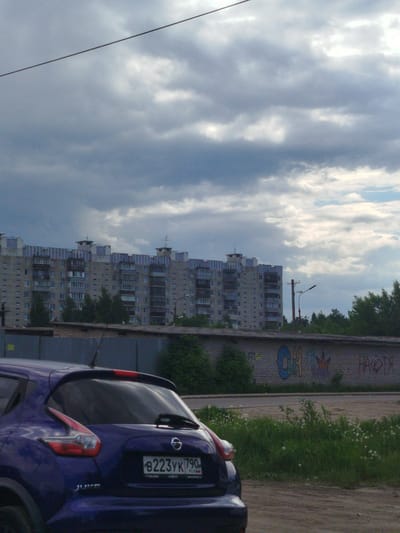 A daytime shot in Noginsk, Russia, features a dark blue Nissan Juke parked on a dirt road. The car's license plate reads "B223YK790". Behind the car is a gray brick wall covered in graffiti, with greenery growing in front of it. Further back is a large, Soviet-era apartment building. The sky is overcast with dark and light gray clouds. A single wire stretches across the upper part of the frame.