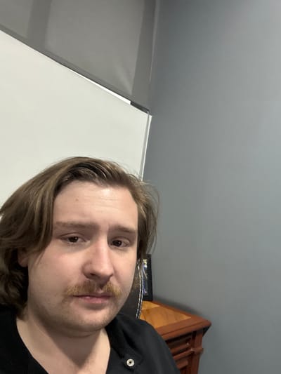 This close-up portrait features a young adult male with medium-length brown hair, a prominent mustache, and light stubble. He looks directly into the camera with a neutral expression. He is wearing a dark top, possibly a shirt or hoodie, with a silver snap button visible near his right shoulder.

The setting appears to be indoors. The upper left portion of the background shows a plain grey ceiling, which meets a vertical white surface (likely a wall or screen) on the left side of the frame. On the upper right, a section of brown wooden furniture, possibly a bed frame or shelving unit, is visible. Below this furniture, a dark, textured fabric is partially seen. The image is lit artificially, and there are no discernible cues for the time of day, weather, or specific location within Cuautitlán Izcalli, Mexico. No text is visible.