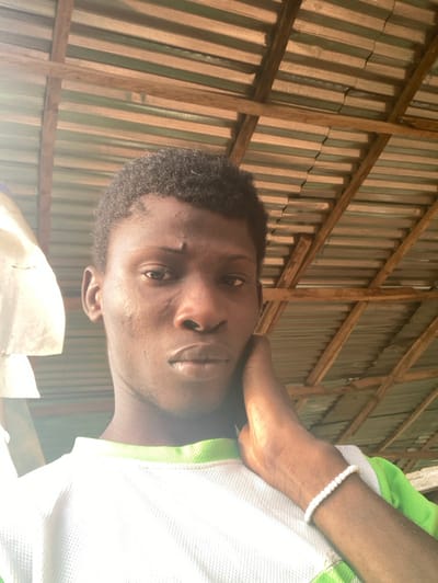 An image depicts a young adult male, likely in Agege, Nigeria, observed from a slightly low angle. The subject, with dark skin and short dark hair, faces forward, looking directly into the camera. A small scar or mark is visible on his forehead above his right eye. He wears a white, mesh-like t-shirt with a light green collar and green accents on the shoulders/sleeves. His left hand is raised, palm against his left cheek and jawline, with fingers slightly splayed. A white beaded bracelet is present on his left wrist. The background features an overhead roof structure composed of wooden rafters supporting corrugated metal sheets, some appearing painted green, creating a striped pattern against unpainted sections. Strong, bright light, originating from the upper left, illuminates the scene, causing glare on the subject's shirt.