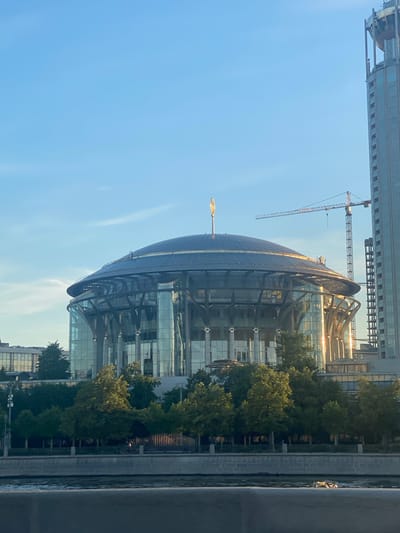 The image depicts a view of a large, circular, glass-fronted building with a domed roof topped with a golden finial, situated in a city. The building appears to be a performance venue or cultural center. Adjacent to it is a tall, modern skyscraper, likely an office building. Construction cranes are visible in the background, suggesting ongoing development in the area. The scene is set under a clear blue sky with some wispy clouds. In the foreground, a body of water, likely a river, is visible, with a stone embankment lining its edge. Trees and greenery are present along the riverbank, creating a contrast between the natural elements and the urban architecture. The lighting suggests it is either early morning or late afternoon, with the sun casting a warm glow on the glass facades of the buildings. There are no people clearly visible in the image, but a lone figure can be faintly discerned on the embankment in the middle ground. The image provides a glimpse of a modern urban landscape with a blend of architectural styles and a touch of nature.