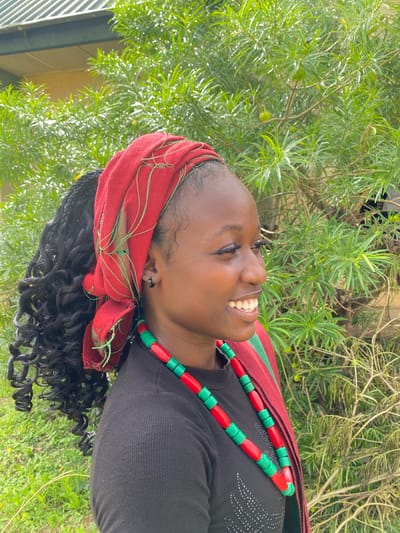 The image shows a young Black woman from the chest up, outdoors. She is wearing a black top, a red and green fabric headscarf, and a necklace made of red and green beads. Her head is tilted back, and she is smiling broadly with her eyes closed. Her hair is dark and curly. The background is filled with lush green foliage, suggesting a natural or garden setting. The lighting indicates it is daytime, likely sunny, with dappled light filtering through the leaves. The woman's expression conveys happiness and contentment. There is no visible text in the image. The location is identified as Wang, Nigeria.