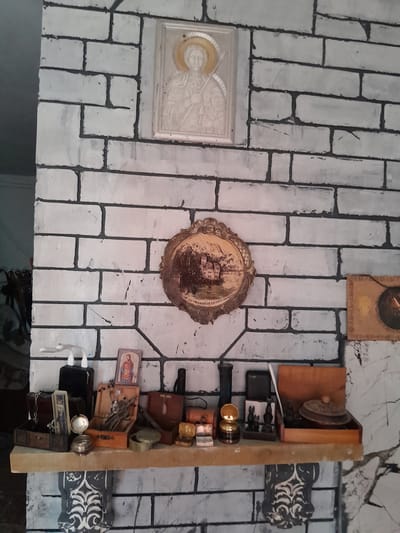 The image displays a wall with a brick-like pattern painted in white with dark grout lines. A shelf is mounted on the wall, filled with various objects. These include several wooden boxes, some open, revealing their contents, such as small statues, trinkets, and what appear to be dried plants or twigs. There is also a small gramophone, a framed religious icon depicting a figure with a halo, and a small, oval decorative plate with a landscape painting. To the right of the shelf, there is a framed picture with a circular object inside. Above the shelf, there are two decorative wall hangings. The lighting suggests an indoor setting, possibly a home or a display area. The objects suggest a collection of decorative and possibly antique items. The location is identified as Cimișlia, Moldova.