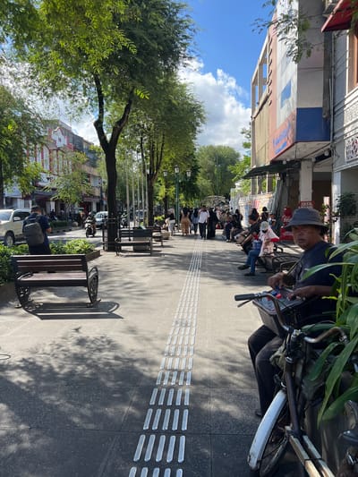 This media file captures a bustling street scene in Yogyakarta, Indonesia, likely during the daytime given the strong sunlight and shadows. The perspective is from a low angle, looking up along a pedestrian walkway lined with trees.
The primary subjects are the people walking and sitting along the sidewalk, interspersed with parked motorcycles and bicycles. A man wearing a hat and dark shirt is prominently featured in the foreground, looking towards the camera while tending to a bicycle. Further down the walkway, several individuals are seated on benches, some with backpacks, suggesting a leisurely stop or waiting period. The architecture of the buildings lining the street is visible, with some showing signs of age and others with colorful facade details. Lush green trees cast dappled shadows on the pavement, creating a pleasant, shaded atmosphere.
The activity taking place is the normal flow of pedestrian and vehicular traffic in a public urban space. People are moving about their day, suggesting a busy marketplace or tourist area. The presence of benches indicates a place for rest and observation.
Notable details include the textured paving stones on the walkway, which appear to be tactile paving for the visually impaired. The sunlight is bright, indicating a clear or partly cloudy day. The overall mood seems relaxed and ordinary, reflecting a typical day in this Indonesian city.
Some visible text can be seen on the building facades, though it is partially obscured and difficult to read in full. One sign appears to have red lettering, possibly indicating a shop or establishment. Another sign reads "JASA" which translates to "SERVICE" in Indonesian. The image provides a glimpse into the vibrant street life and urban environment of Yogyakarta.