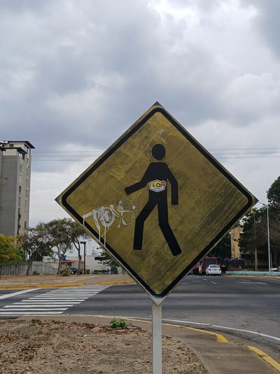 A medium shot of a yellow and black pedestrian crossing sign that has been vandalized with white graffiti. The sign is diamond-shaped and has an image of a person walking in black. A sticker is placed on the person's body, and white graffiti is sprayed at the bottom of the sign. The sign is located near a road with a crosswalk. The location is Guayana City, Venezuela. The sky is cloudy.