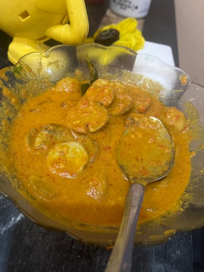 A close-up shot shows a bowl of curry with sliced potatoes and a spoon resting in it. The curry is a vibrant orange-yellow color and appears to be seasoned with chili flakes. The bowl is clear glass with a decorative, wavy rim. In the background, out of focus, is a bright yellow object, possibly a toy, and a white jar. The setting is likely a kitchen or dining area in Beuringen, Indonesia, given the context provided.