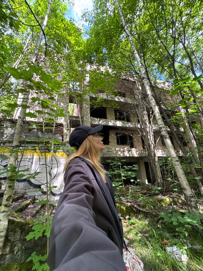 A young woman with blonde hair, wearing a dark baseball cap and a dark jacket over a patterned shirt, stands in a forest looking up at an abandoned brick building. The building is multi-story with visible broken windows and graffiti on the lower levels. Trees, some with birch bark, grow around and through the structure, with dense green foliage obscuring parts of the building and the sky. The ground is covered in leaves, grass, and some scattered debris. The location is the city of Interkompleks, Russia.