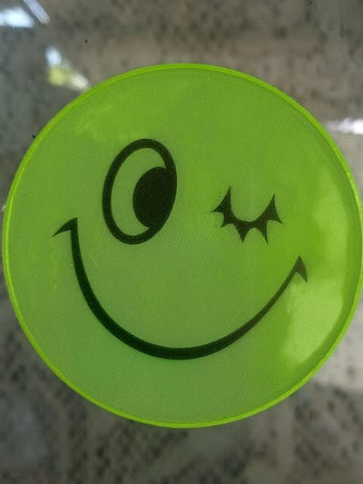 The media file displays a close-up, eye-level, medium shot of a lime green oval sticker featuring a winking smiley face. The sticker is made of fabric with a visible woven texture and has a slightly lighter green border. The smiley face is depicted with a black outline. The left eye is a large circle with a smaller black circle inside, representing a pupil. The right eye is closed, indicated by a stylized, angular shape with three points, resembling a wink. The mouth is a broad, upward-curving smile.

The sticker is affixed to a translucent surface, likely glass, which shows a blurred background of what appears to be outdoor scenery. The background includes muted colors and shapes suggestive of natural elements, possibly foliage or pavement, with some reflections indicating light sources. The overall impression is bright and cheerful due to the vibrant green of the sticker and the smiley face design.

There is no visible text in the media file. The image does not depict any people or specific activities. The lighting suggests it could be daytime, and the reflections hint at an outdoor or well-lit indoor location. The setting is not definitively identifiable beyond being attached to a surface with a view of a blurred exterior. The emotion conveyed is one of playfulness and good humor, characteristic of a smiley face. The provided location context of Stavchyntsi, Ukraine, is not visually discernible from the image itself.