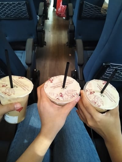 Two individuals are seated in a vehicle, likely on a bus or train, holding drinks with straws. The seats are upholstered in dark blue fabric, and the floor appears to be wood laminate. The drinks are in clear plastic cups with white lids featuring a coffee bean illustration and text in a language that is likely Indonesian, with phrases like "1000 Stores Worldwide" and "Dari Indonesia". One person's arm is visible, wearing blue denim jeans. The context of Karawang, Indonesia, suggests this might be a travel scene within or to/from that city. The lighting indicates an indoor environment, possibly during daylight hours given the ambient light.