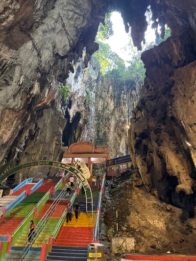 The image presents a grand view inside a massive natural limestone cave, characteristic of the Batu Caves located within the Selayang Municipal Council, Malaysia. The main focus is a long, multi-colored staircase, painted in bright bands of red, orange, yellow, green, blue, black, and gray, ascending through the cavernous space. White handrails run along the steps.

Several people are visible on the staircase, appearing to be walking up or down. They are dressed in casual clothing, suggesting they are visitors or pilgrims exploring the site. Near the top of the stairs, an elaborate golden-green archway adorned with intricate designs and text in Tamil script stands prominently. Behind this archway, structures hinting at a temple complex are nestled within the cave.

The cave walls and ceiling are rugged, showcasing natural rock formations and textures. A large opening towards the top allows abundant natural light to stream in, illuminating the scene and revealing lush green trees and a bright sky outside, indicating it is daytime. On the right side, blue banners bear text in Tamil script and English, which reads "SRI VALLI DEIVANAI." A small box labeled "DONATION BOX" is also visible beside the stairs. The scene conveys a sense of wonder at both nature's grandeur and human devotion, as people traverse this unique and colorful pathway.