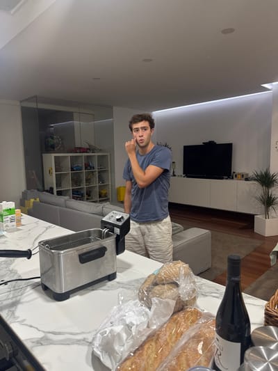 The image depicts an interior scene within a modern residential kitchen and living area in Cidade da Maia, Portugal. In the foreground, a white marble-patterned countertop holds a stainless steel deep fryer with a visible basket, several plastic-wrapped bread items (baguettes and rolls), and a dark glass bottle, possibly wine, with "JAVALI R" visible on its label. A white carton and another small container are present behind the fryer. Standing behind the countertop is a male subject, appearing young adult, with light brown hair, wearing a dark blue t-shirt and light-colored shorts. He faces slightly right of the camera, looking directly forward with an open mouth, his right index finger placed near his right temple.
The background shows a contemporary living space with a large grey sectional sofa. To the left, a transparent glass partition fronts a white shelving unit displaying various model vehicles and what appear to be yellow construction blocks. To the right, a large flat-screen television is mounted above a long white floating media console. A tall green potted plant in a white square planter stands to the far right. The room features white walls, recessed ceiling lighting, and linear ambient strip lighting along the top of a wall unit. The flooring in the living area is polished wood.