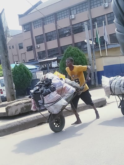 A young Black male, wearing a yellow t-shirt and black shorts, is pushing a loaded wheelbarrow down a street in Shangisha, Nigeria. The wheelbarrow is filled with various bags and bundles of goods. He is barefoot and his body is tense with exertion. In the background, a modern building with a blue and yellow facade is visible, along with a fence and some trees. Several flags, including a green one and a grey one, are planted in front of the building. The street appears to be paved and there is a concrete curb along the side. Another wheelbarrow, also laden with goods, is partially visible to the right. The sky is overcast, and the overall lighting suggests it is daytime.