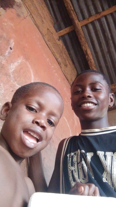 Two African boys smile for a selfie. The younger boy, on the left, has his mouth open and is missing a front tooth. The older boy, on the right, wears a black basketball jersey with white lettering. They stand against a background of an orange wall and a corrugated metal roof with wooden beams. The lighting is bright, casting soft shadows on their faces.