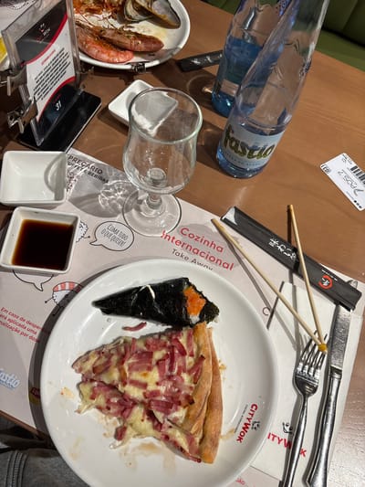 The image depicts a dining table scene, likely in a restaurant located in Vila Nova de Gaia, Portugal, identifiable from the "Fastio" water bottle, a brand of water from Serra do Gerês, Portugal. The table is cluttered with various items, including a bottle of water, an empty wine glass, a plate with a piece of pizza and a Temaki sushi, a plate of seafood (shrimp and mussels), chopsticks in a paper wrapper, cutlery (knife and fork), small square dishes (one empty and one with soy sauce), menus, and a table card with the number 31. The restaurant branding "City Wok - Cozinha Internacional" is visible on the plate and menu, suggesting an Asian-themed cuisine. The presence of a bill/receipt and used dishes indicates that a meal is in progress or has recently concluded. The environment is indoors, with the table surface appearing to be made of wood.