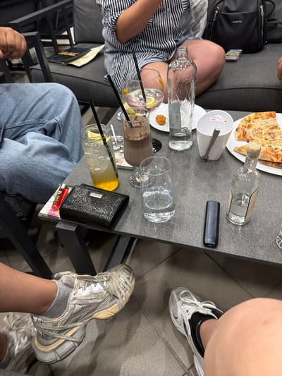 A group of people are gathered around a table at an outdoor cafe in Sapareva Banya, Bulgaria. On the table, there are several drinks including a glass of yellow liquid with ice and a lemon slice, a glass of chocolate milkshake, and an empty glass. There is also a slice of pizza, a small bowl of what appears to be a fried snack, a clear bottle of water, and a bottle of clear liquid. A black wallet, a red lighter, and a black electronic device are also visible on the table. Two individuals' legs and feet are visible, wearing casual clothing and sneakers. In the background, a black sofa and a black backpack can be seen. The setting suggests a relaxed, social gathering.