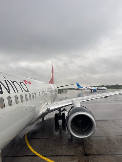 The image shows a view from inside an airplane at Bolshoye Savino Airport in Perm, Russia. The wing and engine of the airplane are visible, along with another plane in the distance. The ground is wet, suggesting it has been raining. The sky is overcast. The name "wing" is visible on the side of the airplane.
