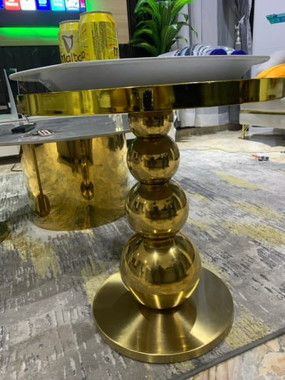 A golden candelabra, composed of a circular base, three spherical joints, and a circular top, stands prominently. A slender metallic structure with a gray top plate ascends vertically from the candelabra's top. In the background, a can of Malta beverage and a green plant are visible. The candelabra rests on a patterned gray rug.