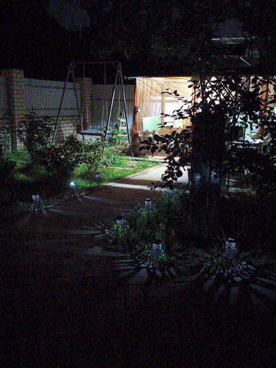 The image depicts a dark backyard scene in Kotovsk, Russia. In the background, a brick building with a light-colored tarp or sheeting extends across it, with visible windows and interior lighting suggesting a covered patio or an extension to a house. A swing set with metal A-frame supports and wooden seats is present in the mid-ground, situated near the building. The foreground is characterized by grass, bushes, and a paved pathway or patio. Several small, decorative solar-powered lanterns are placed along the pathway and in the grass, casting patterned light and shadows on the ground. The overall atmosphere is nocturnal and appears to be residential.
