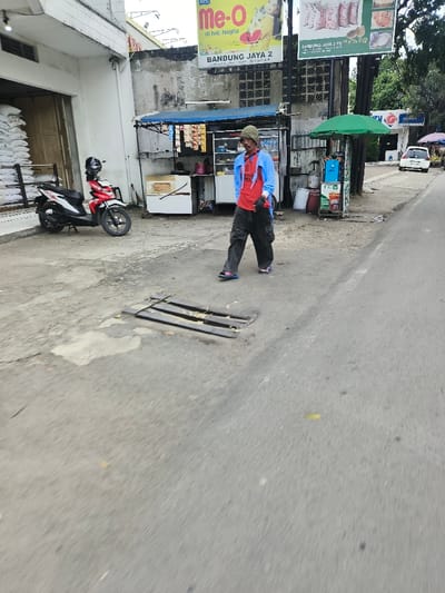 A man in a red shirt, blue jacket, and loose black pants walks on a street. A metal grate is visible in the foreground. Behind him, there is a small food stand with a blue awning. A parked red and white scooter is to the left, next to a white building. A green umbrella is beside the stand. Various signs and billboards are displayed above. A white car is parked further down the road.