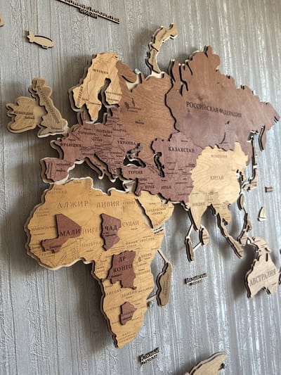 The image displays a wooden world map mounted on a textured grey wall. The map is composed of individual wooden pieces representing continents and countries, with some cities and geographical features labeled in Russian. Several small wooden airplanes are attached to the wall near the map. The lighting suggests an indoor environment. The overall scene appears to be a decorative display. No specific location in the city of Лесные Поляны, Russia is identifiable from the image itself, as it is a generic interior shot of a world map.