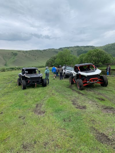 The image depicts an outdoor scene with three off-road vehicles, two of which are prominently displayed in the foreground. The vehicle on the left is black with red accents, and the vehicle on the right is white with red accents. Both vehicles appear to be UTVs (Utility Terrain Vehicles) or similar recreational off-road vehicles. In the background, a third, lighter-colored off-road vehicle is partially visible.

Several people are present in the scene, some standing near the vehicles and others walking. One person in the foreground, near the black vehicle, is wearing a teal jacket and dark pants, with a helmet on. Other individuals are dressed in casual outdoor attire.

The environment is a grassy, uneven terrain with muddy patches and tire tracks indicating recent vehicle activity. In the distance, rolling hills covered in greenery and scattered trees rise under a cloudy sky. A body of water, possibly a river or a stream, is visible in the far right of the image, with a few individuals standing near its edge. The overall impression is a group engaged in recreational off-roading or an outdoor adventure. The reference to "Yelansy, Russia" does not correspond to any recognizable geographical features in the image, suggesting it may be extraneous information.