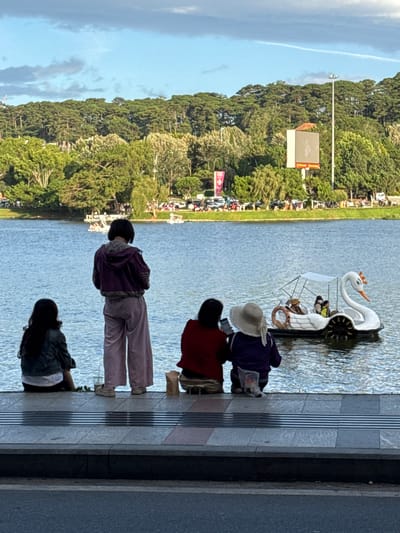 In Xuan Huong Lake, Da Lat, Vietnam, four people sit on the edge of the promenade, facing the water. In the background, a swan-shaped paddle boat with several occupants glides across the lake. Beyond the lake, a tree-lined hill rises, topped by a large billboard and a tall light pole. The sky is a mix of blue and gray clouds. The water reflects the sky and the surrounding greenery. The foreground shows the paved promenade and a street. The overall scene suggests a peaceful afternoon at a popular tourist spot.