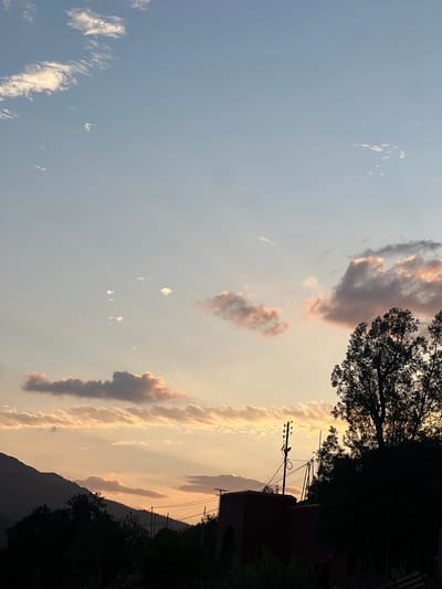 The image depicts a sky scene at sunset in Bin El Ouidane, Morocco. The sky is a mix of pale blue and orange hues, with elongated clouds reflecting the setting sun. Some clouds have a light pinkish tint. Silhouetted trees and a building partially obstruct the view on the right side of the frame. Electrical poles and wires are visible against the colorful sky. The lower part of the frame shows a dark, mountainous landscape.